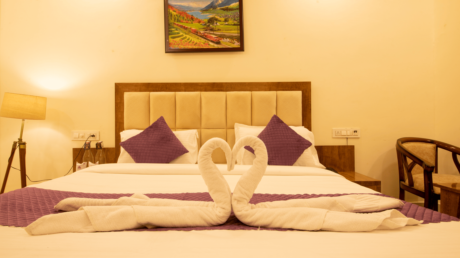 A hotel room, featuring a large bed with a towel swan display, a purple runner and pillows, a wooden headboard, and a painting above the bed | Perfectstayz Value Hills, Rishikesh