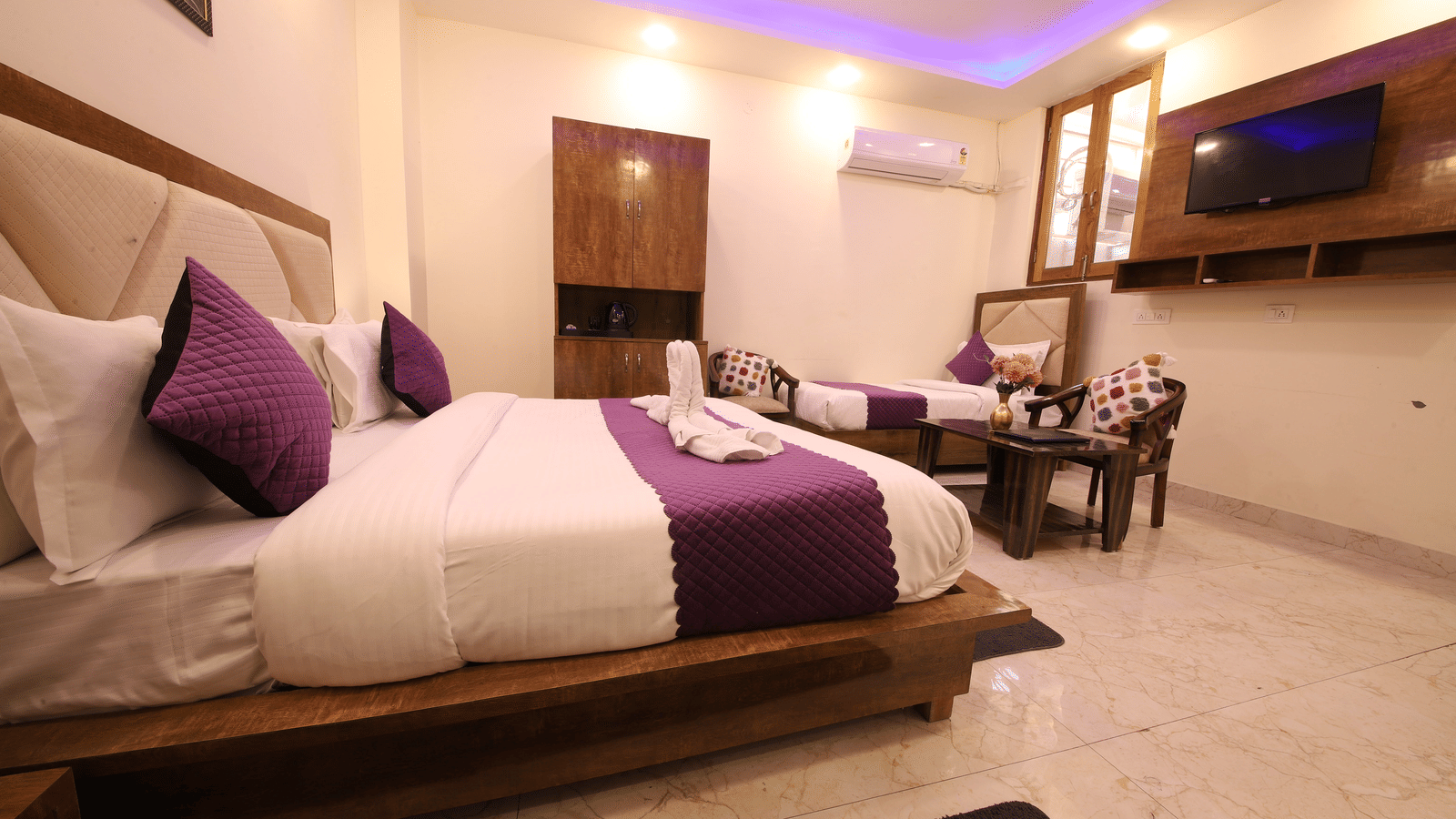 A hotel room with purple cove lighting, featuring a large bed with white linens and purple runner, a wall-mounted TV, and a seating area | Perfectstayz Value Hills, Rishikesh