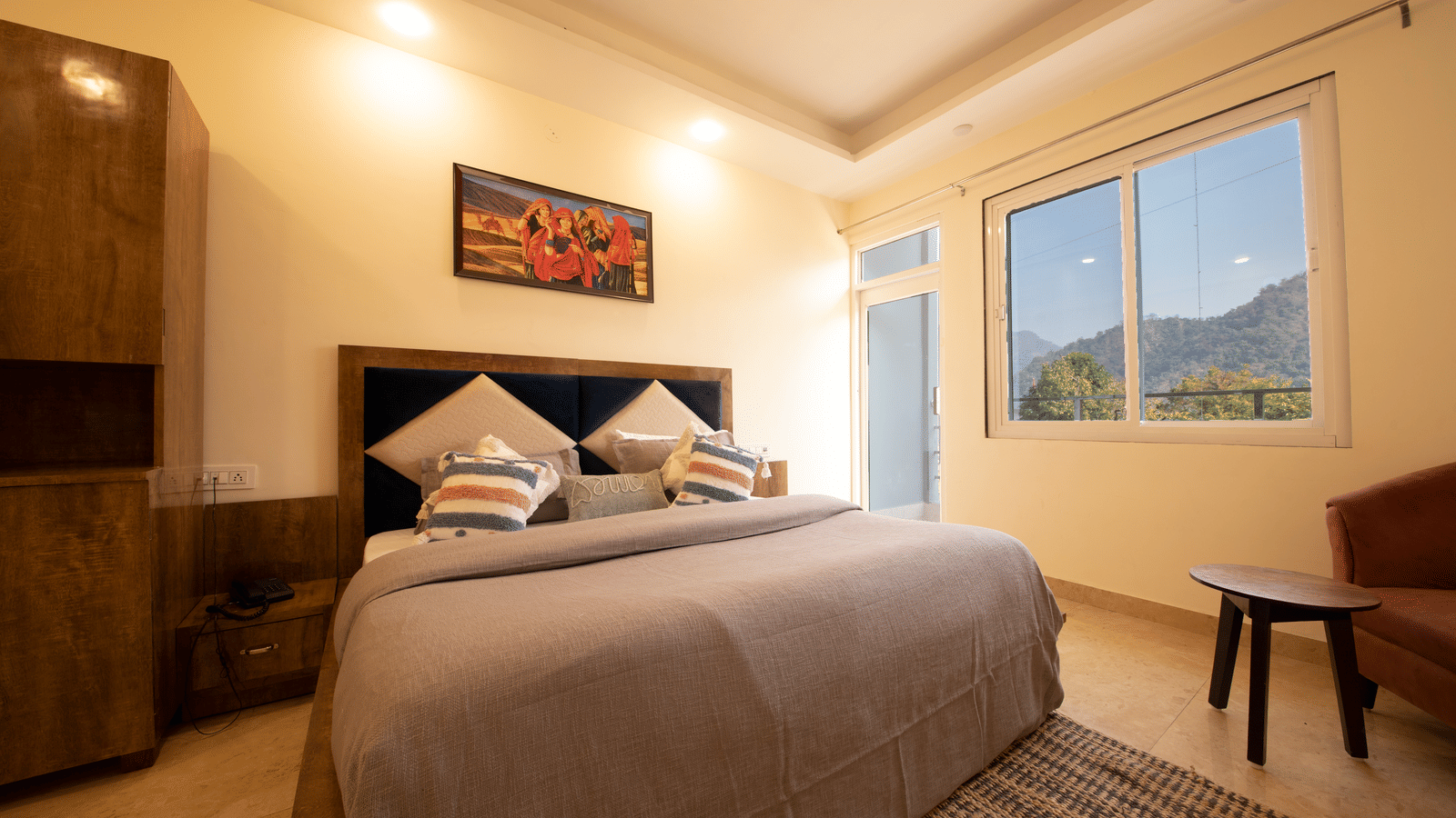 A hotel room, featuring a large bed with a grey duvet and a wooden frame, a painting on the wall, and a window with a view of hills | Perfectstayz Value Hills