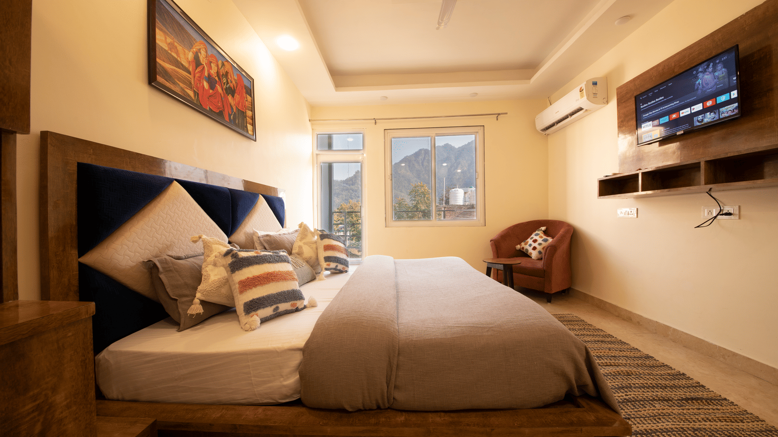 A hotel room, featuring a large grey bed with white linens, blue and orange pillows, a wooden frame, a wall-mounted TV, and a large window with white curtains | Perfectstayz Value Hills, Rishikesh