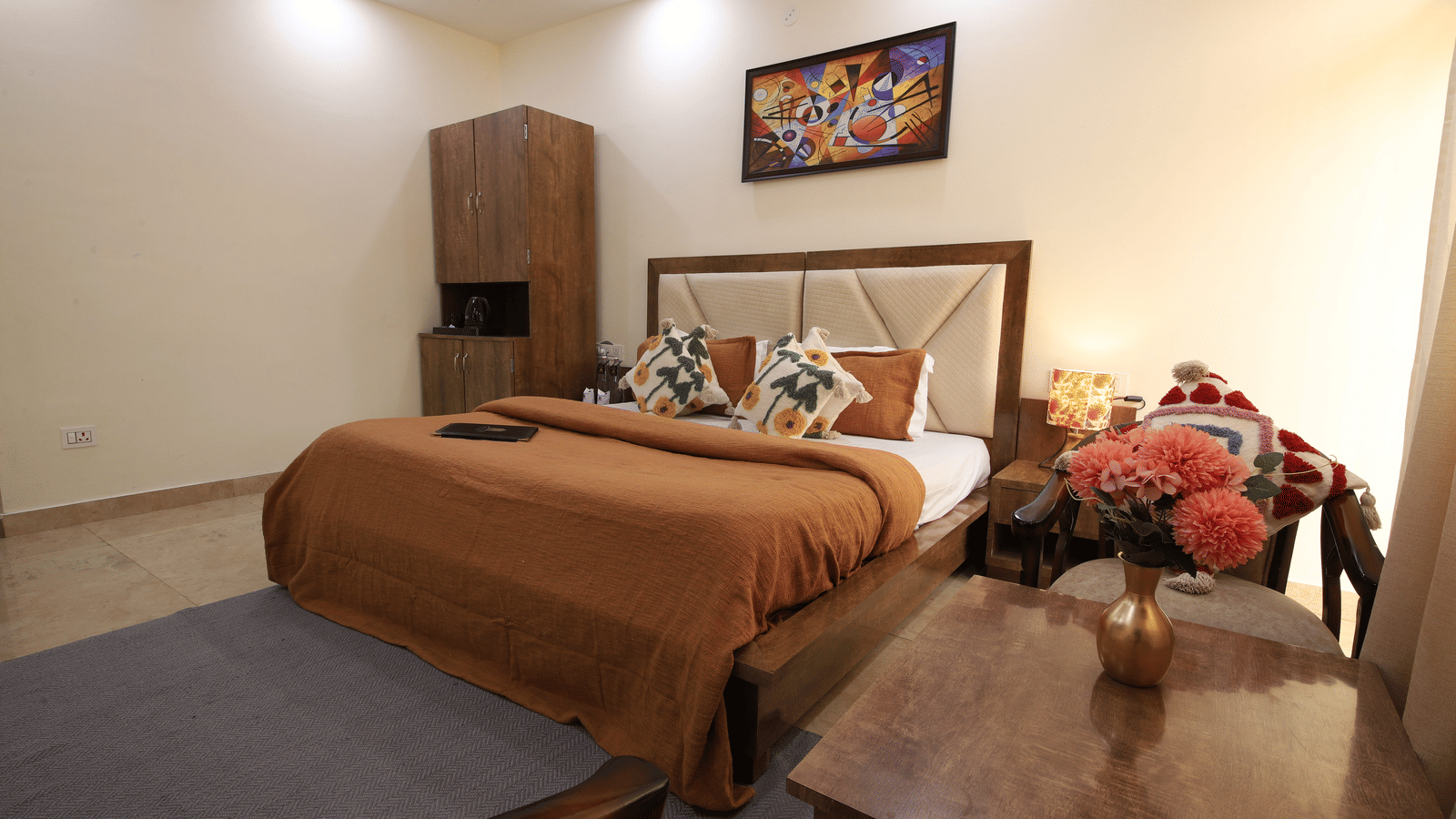 A hotel room, featuring a large bed with a brown duvet, a wooden nightstand in the foreground, and a colourful painting above the bed | Perfectstayz Value Hills, Rishikesh