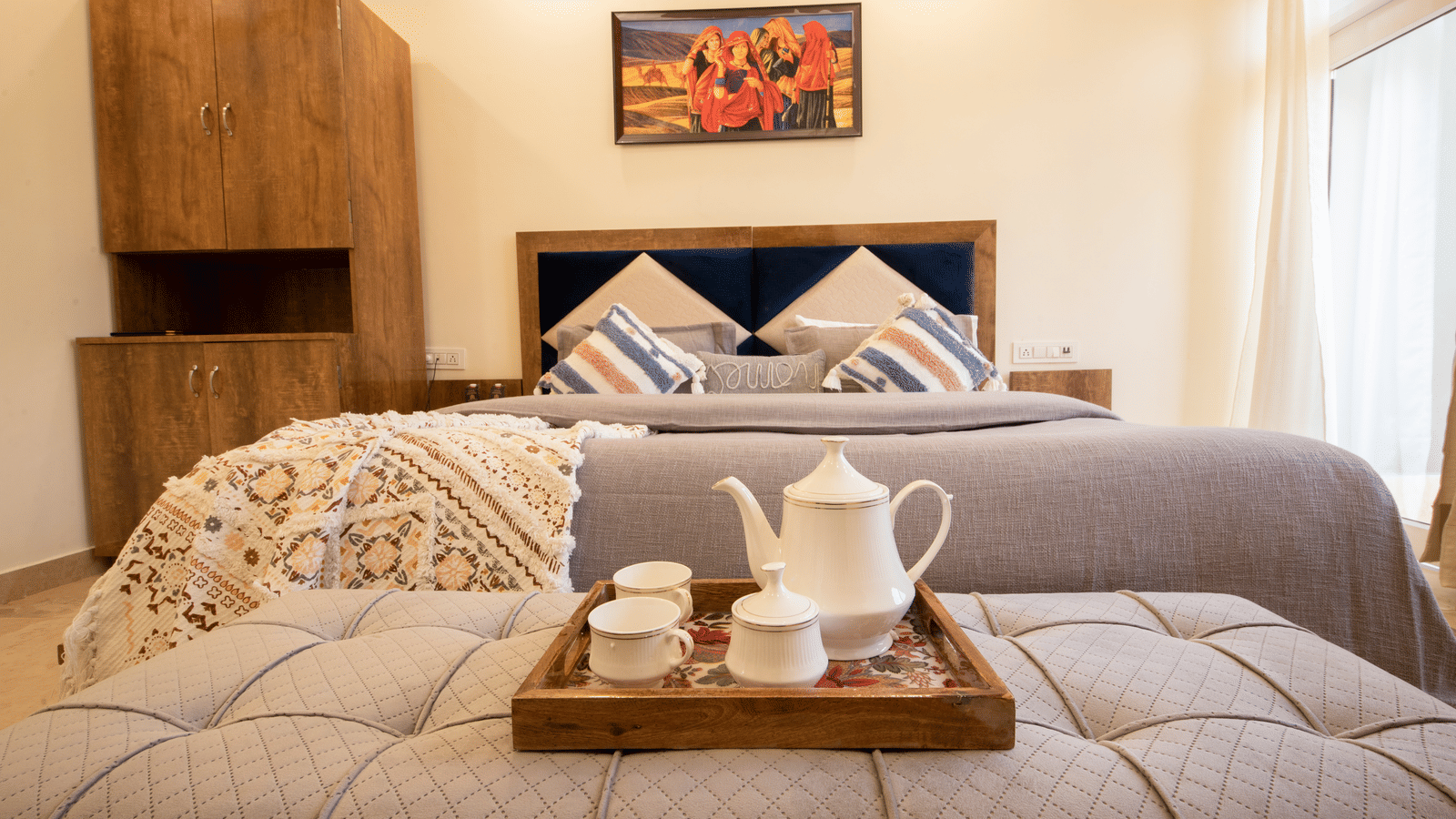 A hotel room, featuring a large grey bed with white linens, blue and orange pillows, a wooden tray with a teapot and cups on the bed, and a painting on the wall | Perfectstayz Value Hills, Rishikesh