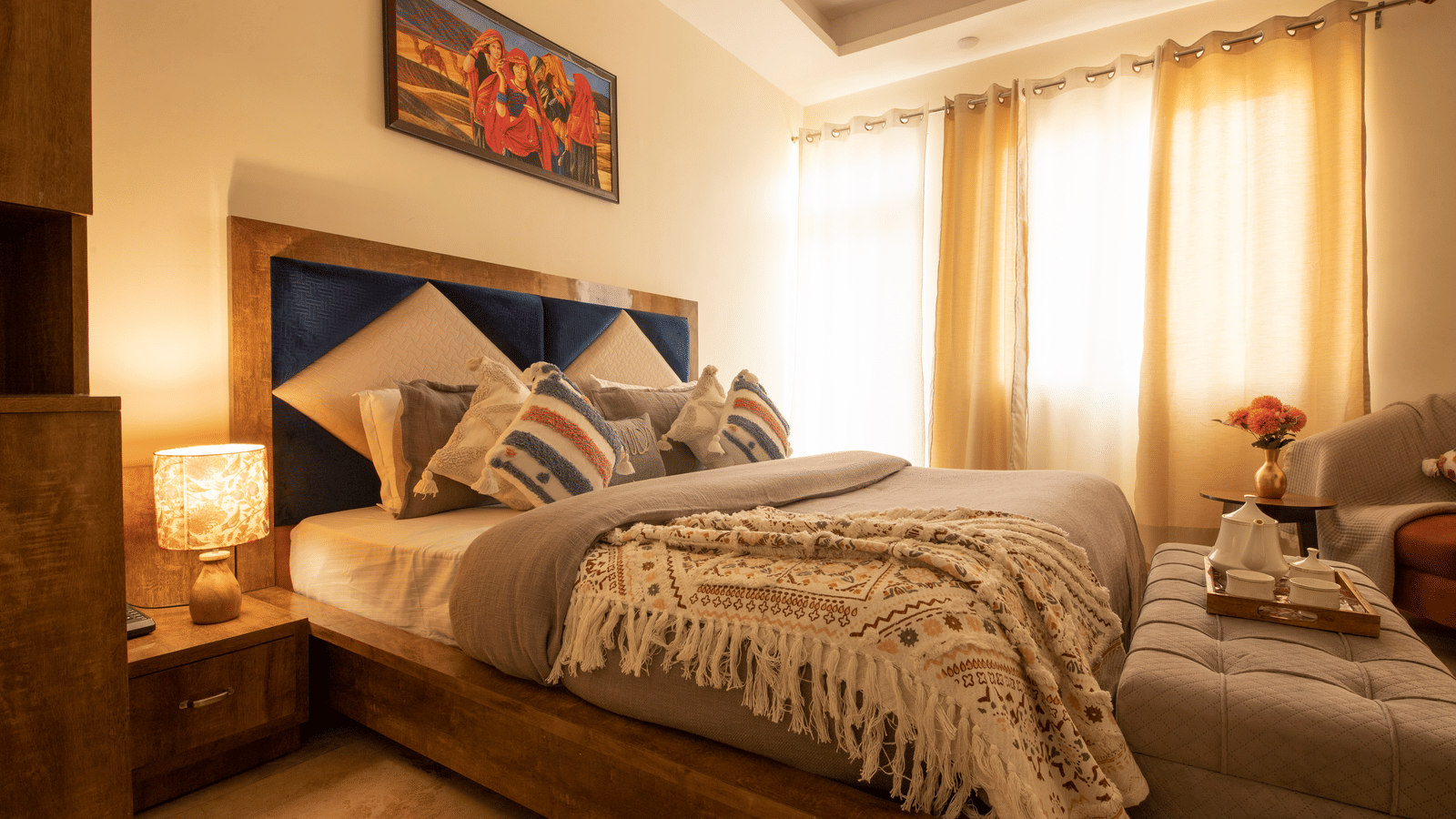 A brightly lit hotel room, featuring a large bed with a wooden frame, multiple decorative pillows, a knit throw blanket, and a window with sheer curtains | Perfectstayz Value Hills