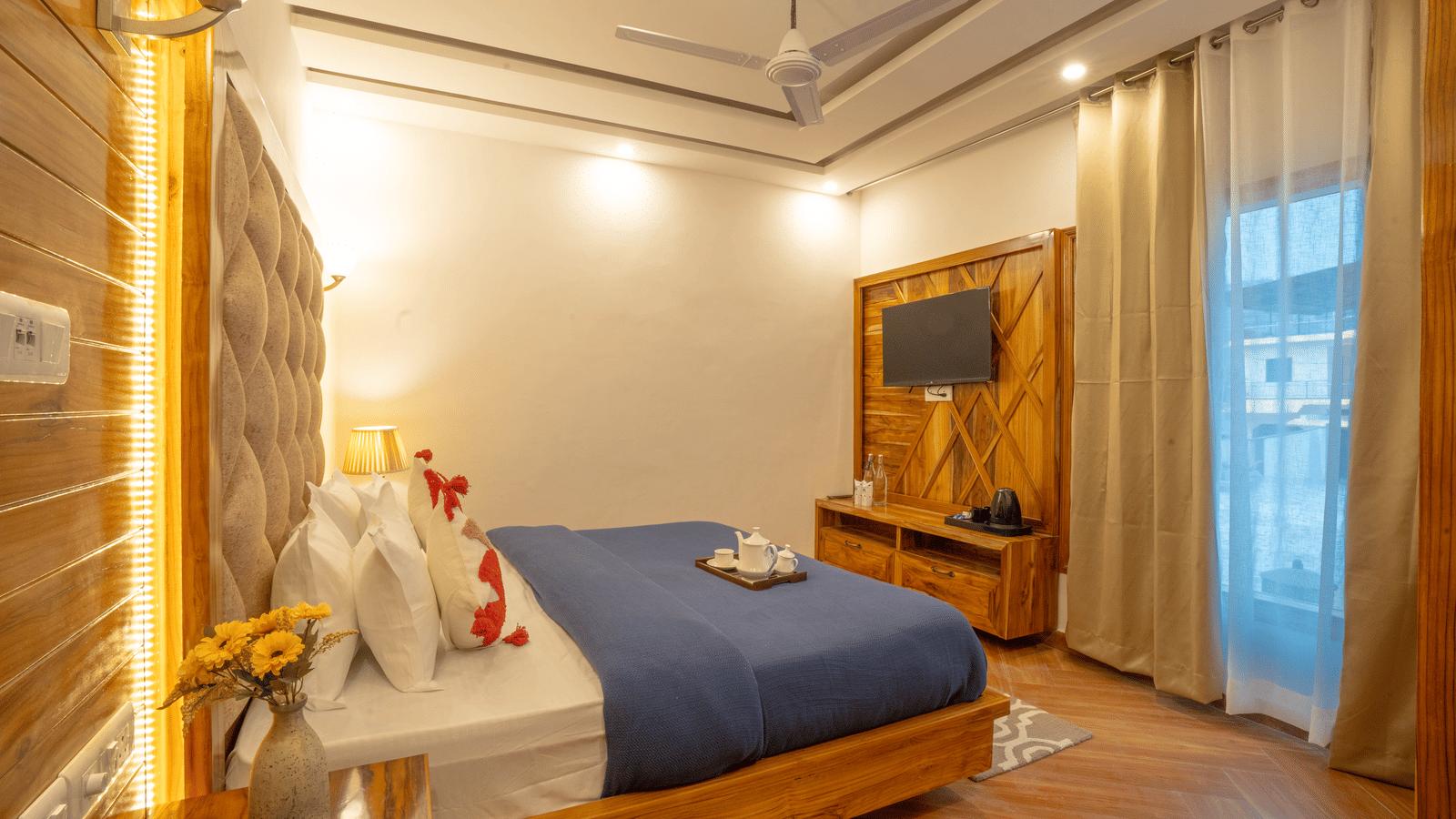 A bedroom with a king size bed, a wall-mounted television, a bedside table, a lamp, and a cabinet at Perfectstayz Value Paradise, Mussoorie.