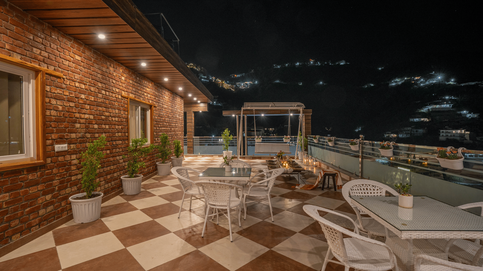 An outdoor terrace with checkered flooring, tables and chairs, potted plants, and hillside night views at Perfectstayz Value Paradise, Mussoorie.
