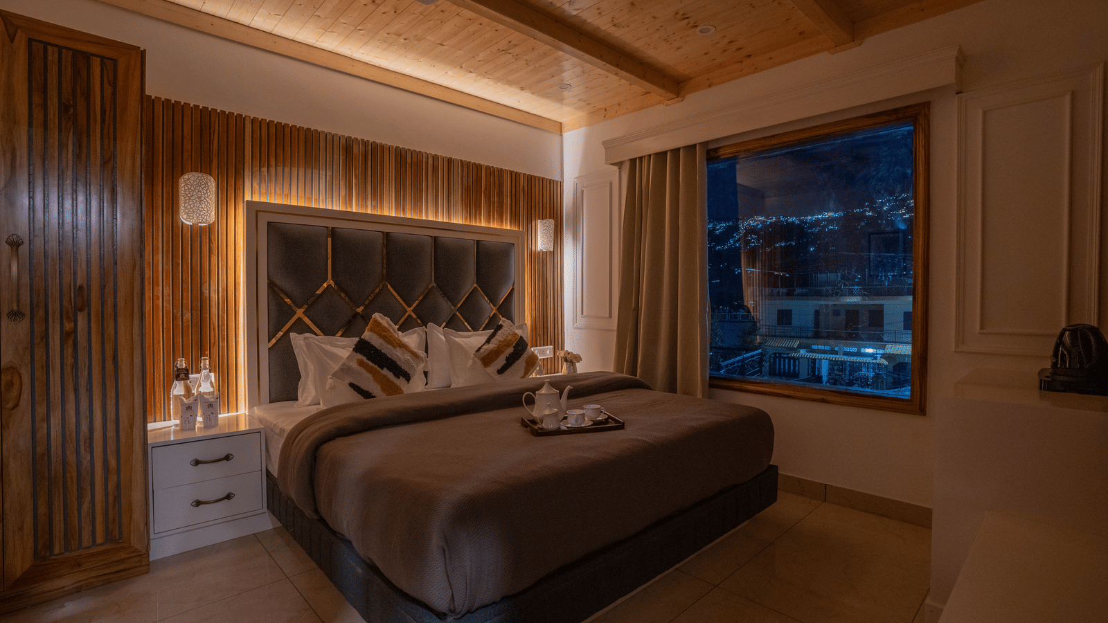 A bedroom featuring a king size bed, a bedside table, a tray with cups and a kettle, ceiling panels, and a window offering a city view at Perfectstayz Value Paradise, Mussoorie.