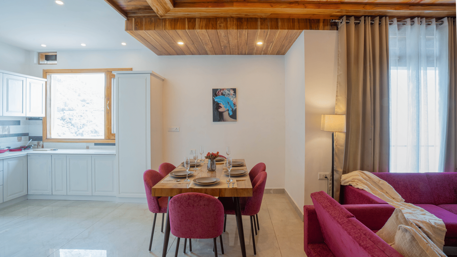 A dining area with a dining table with plates and glasses, chairs, a kitchenette in the background, 2 sofas and large windows at Perfectstayz Value Paradise, Mussoorie.