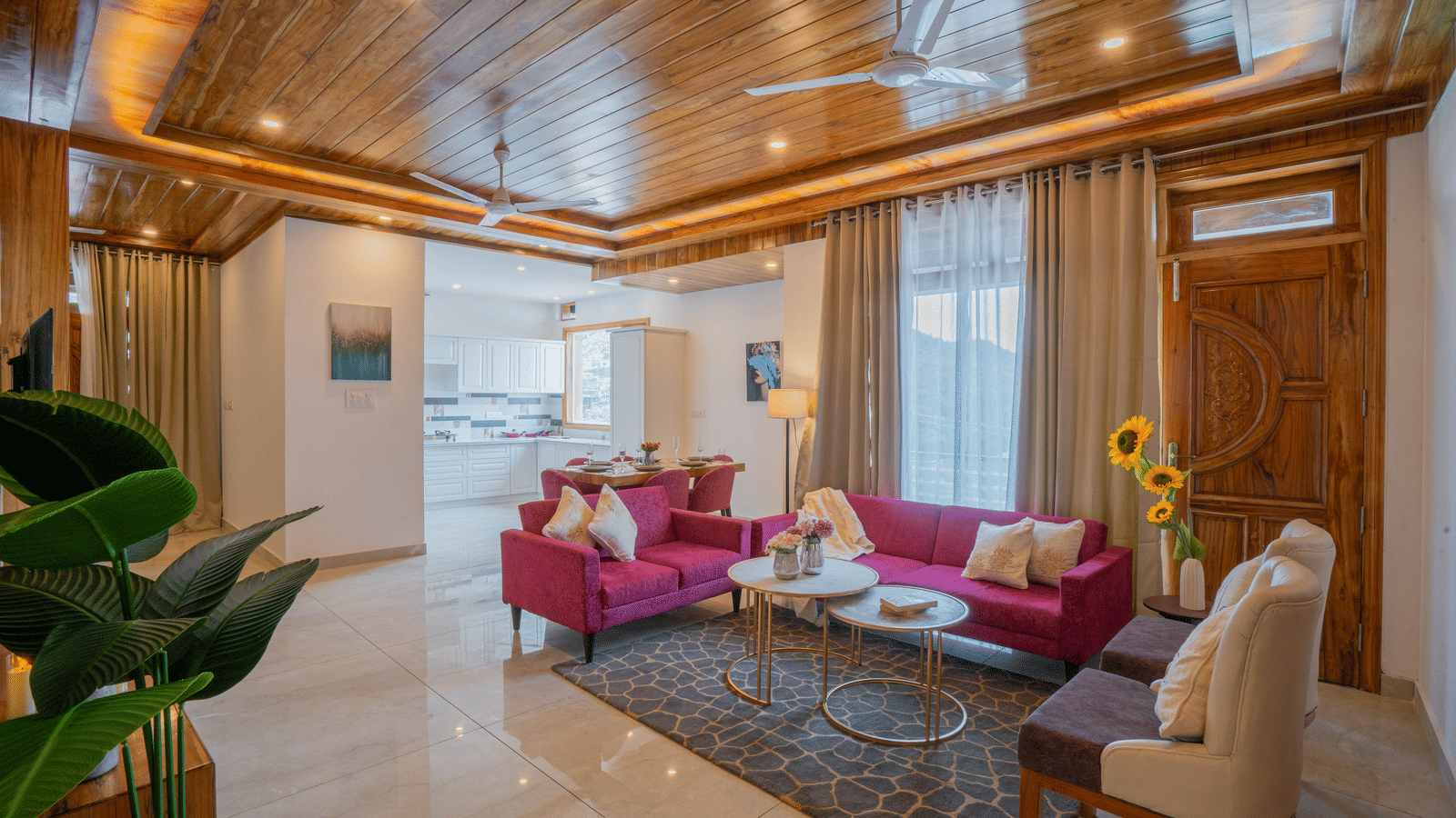 A living room with 2 sofas, a chair, 2 tables, a plant, a wooden ceiling, a dining area, and large windows with curtains at Perfectstayz Value Paradise, Mussoorie.