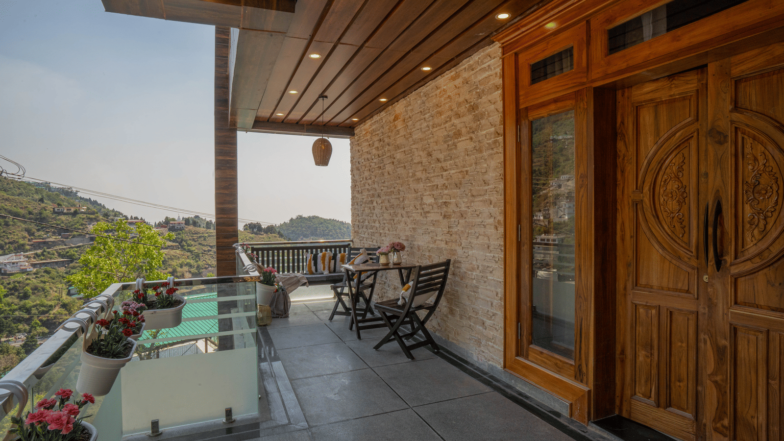 A balcony with a table, a bench, pillows, chairs, and potted plants overlooking hills at Perfectstayz Value Paradise, Mussoorie.