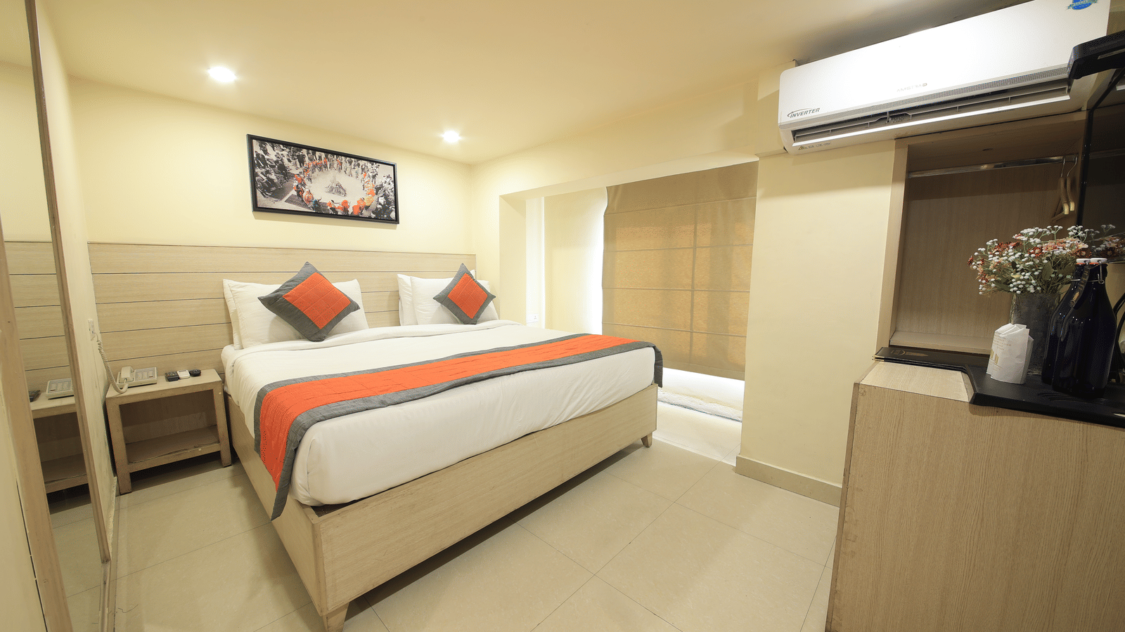A hotel room, featuring a double bed with an orange accent runner, a dark wood wardrobe, and a corner display with flowers | Perfectstayz Value Sagar