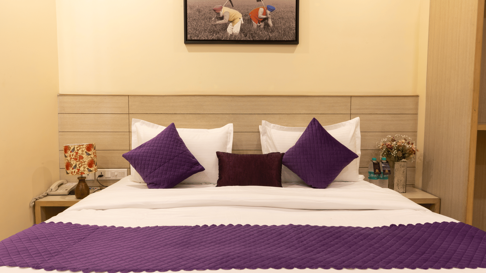 A close-up of a large bed, featuring a white duvet, purple runner and pillows, a light wooden headboard, and a small framed painting on the wall | Perfectstayz Value Sagar