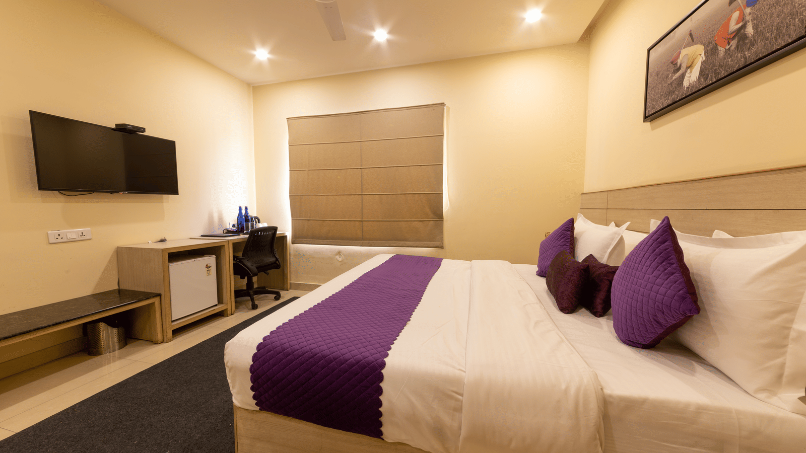 A hotel room, featuring a large bed with a purple runner and pillows, a ceiling fan, a wall-mounted TV, and a desk area | Perfectstayz Value Sagar