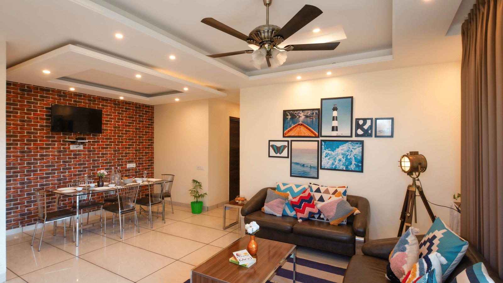 A living area at Perfectstayz Value Shimla (Namah Retreat) with a sofa, wall art, television, dining table, chairs, ceiling fan, and brick-patterned wall.