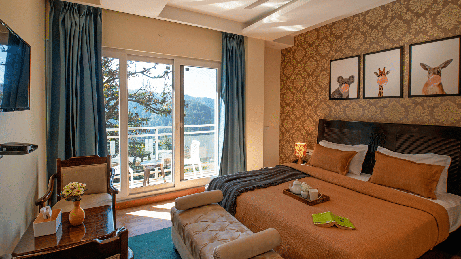 A bedroom at Perfectstayz Value Shimla (Namah Retreat) with a king size bed, wall art, a television, a table, 2 chairs, a daybed, a tray with cups and a kettle, and a sliding door leading to a balcony with a table and chairs.