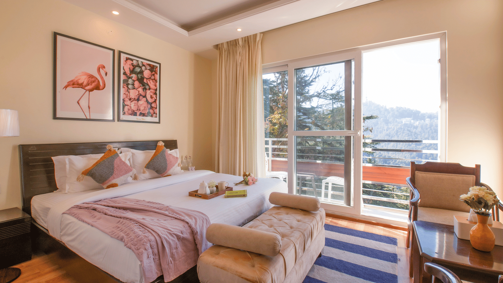 A bedroom at Perfectstayz Value Shimla (Namah Retreat) with a king size bed with a tray of cups and a kettle, wall art, a table, 2 chairs, and large glass sliding doors opening to a balcony.