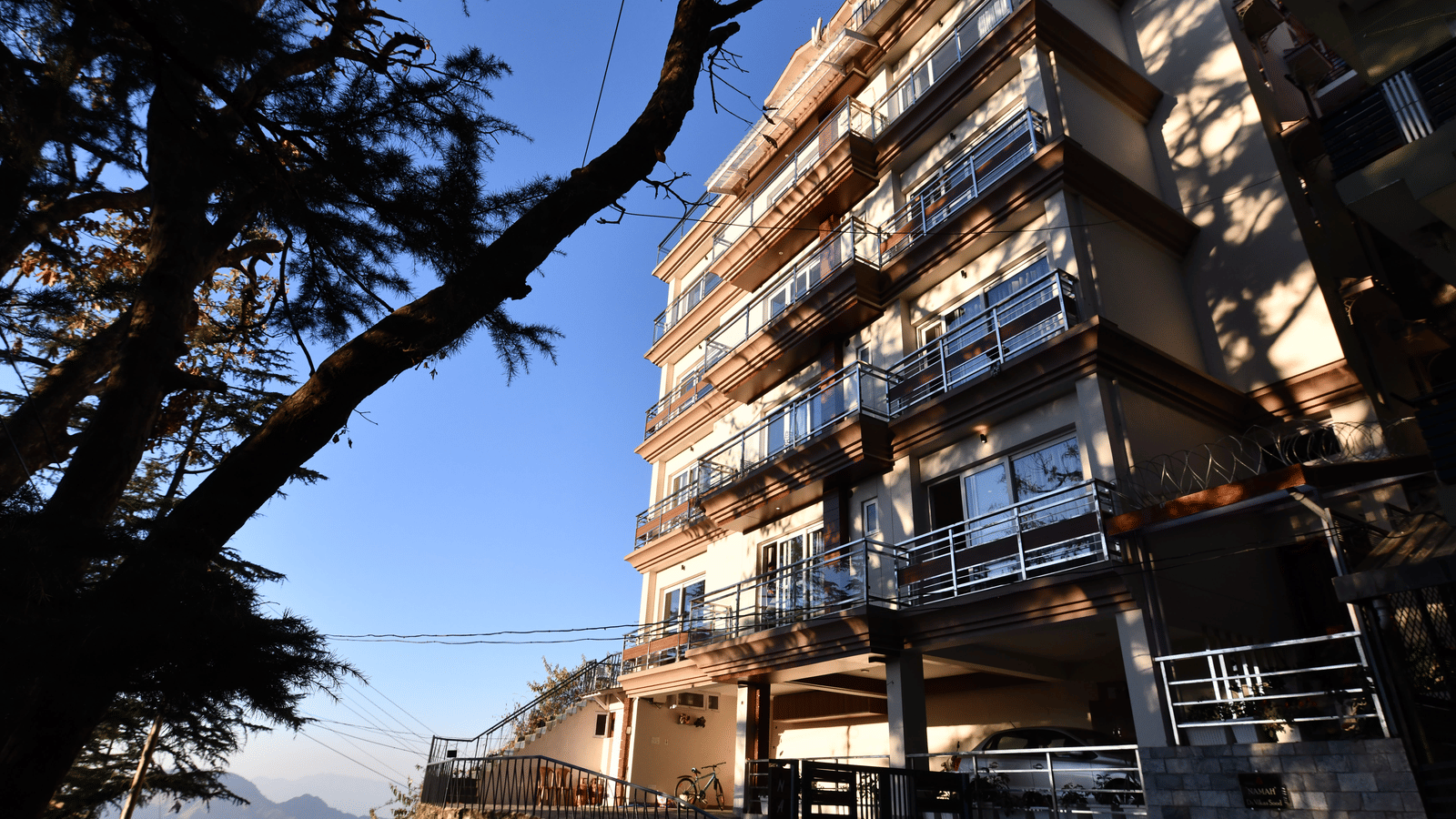 A facade of Perfectstayz Value Shimla (Namah Retreat) with balconies viewed from below against a clear sky and a tree in foreground.