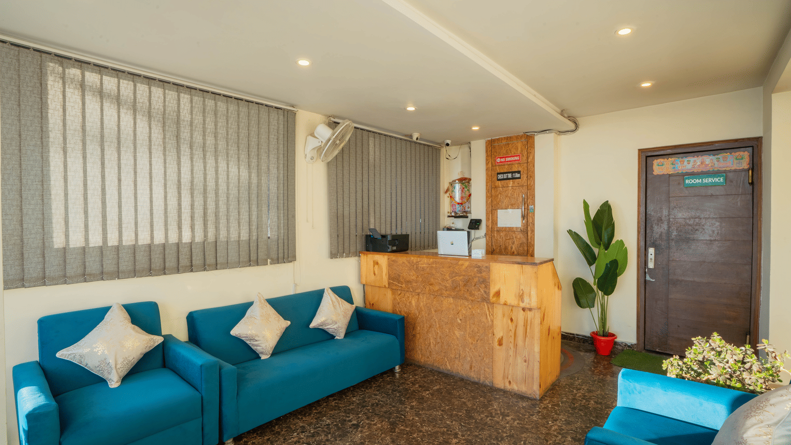 A hotel lobby or waiting area, featuring teal blue sofas and a reception desk in the background, with vertical blinds on the windows | Perfectstayz Value Valley Vista