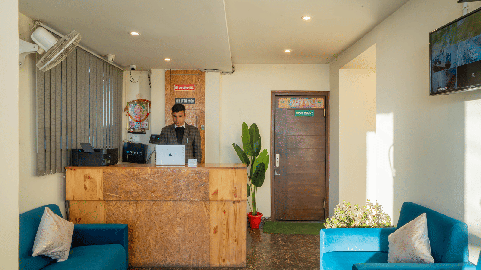 A hotel reception area, featuring a light wood desk, a staff member sitting behind it, and a waiting area with teal blue sofas | Perfectstayz Value Valley Vista