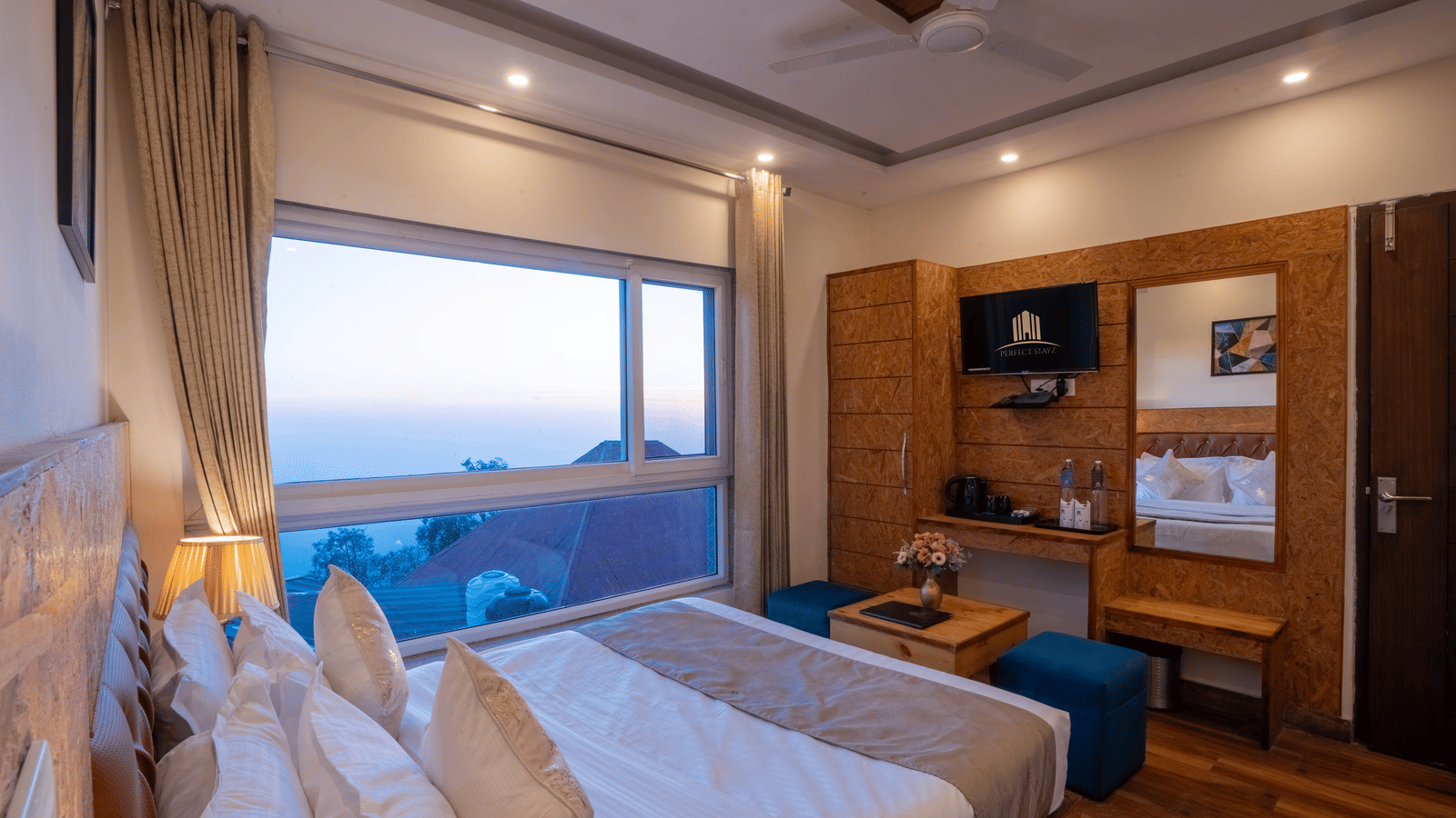 A hotel room with a large window view, featuring a bed with white linens, a blue sofa, a ceiling fan, and wooden furniture | Perfectstayz Value Valley Vista