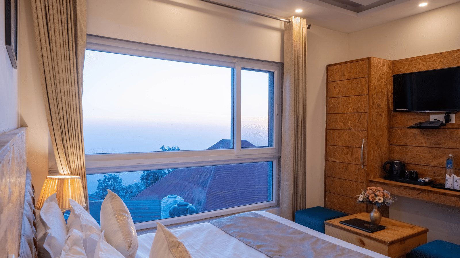 A hotel room with a large window view of a hilly landscape, featuring a bed with white linens and a desk beneath a wall-mounted TV | Perfectstayz Value Valley Vista