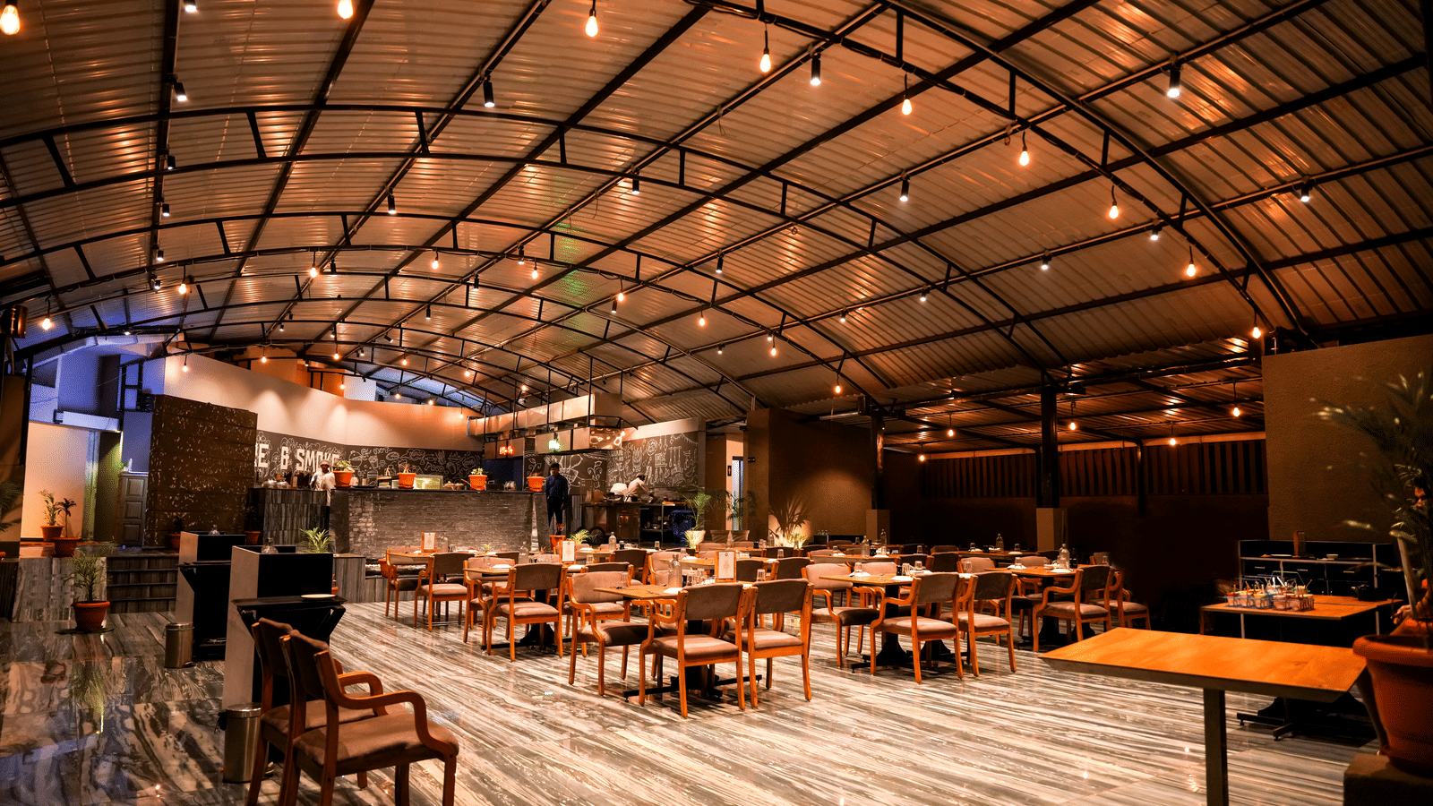  A wide view of the spacious dining hall with wooden tables and chairs under a curved, industrial-style roof at Pravasa Hotel, Kolhapur.