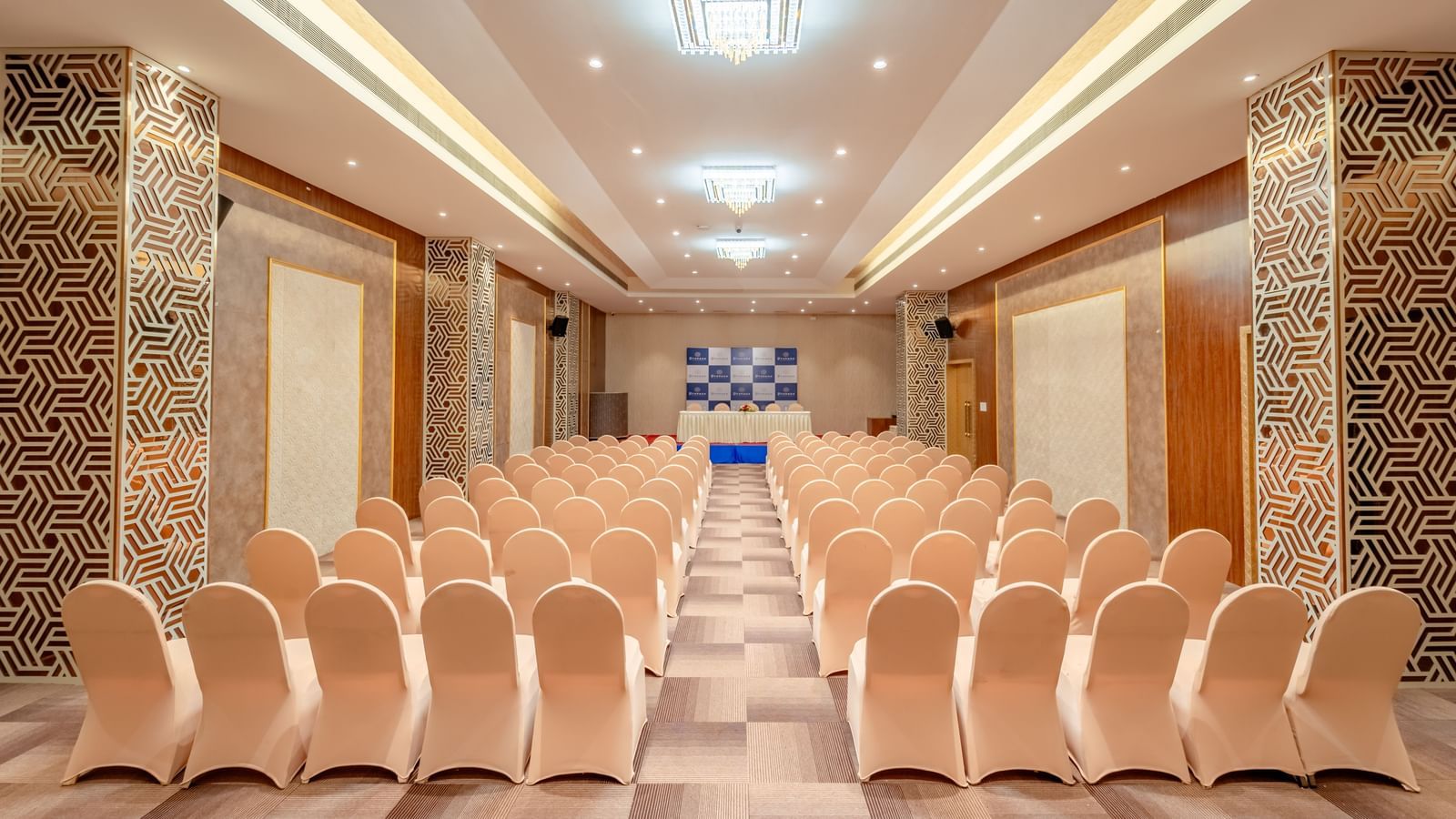 An empty event space features multiple rows of chairs and a prominent stage area | Meetings & Events | Pravasa Hotel, Vijayapura