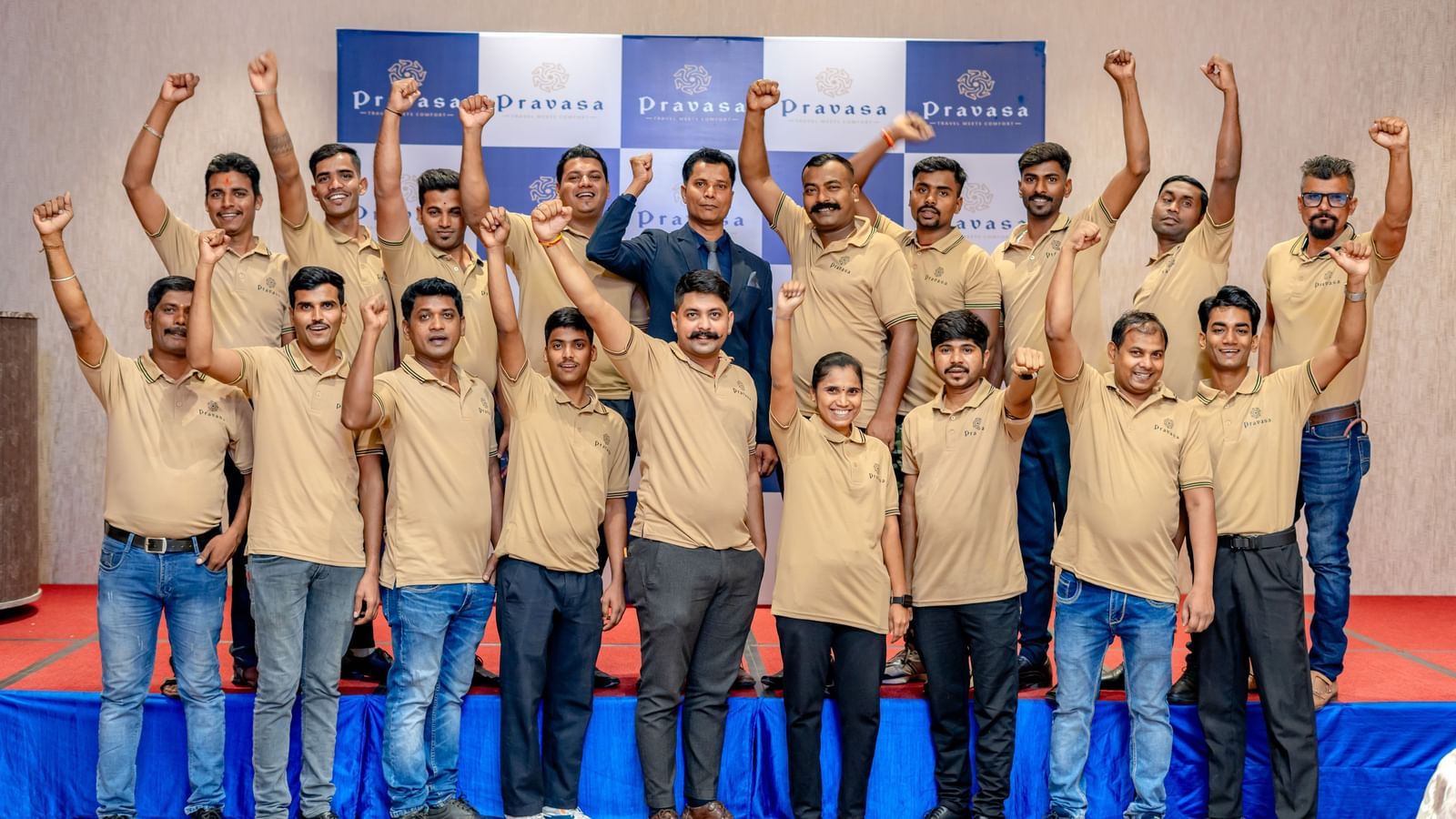  A large group of men in matching tan shirts and blue jeans stand with their arms raised in celebration in front of a blue and white backdrop.