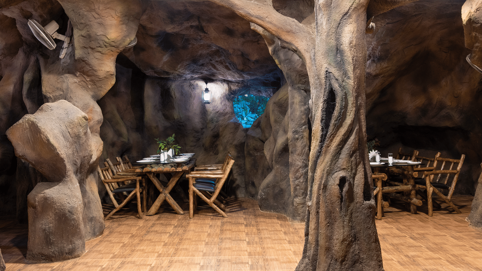 A cave-themed restaurant shows more seating arrangements with wooden tables and chairs amidst sculpted rock walls inside Cave Restaurant at RD's Nature Retreat - The best place for a One Day Trip From Bangalore.