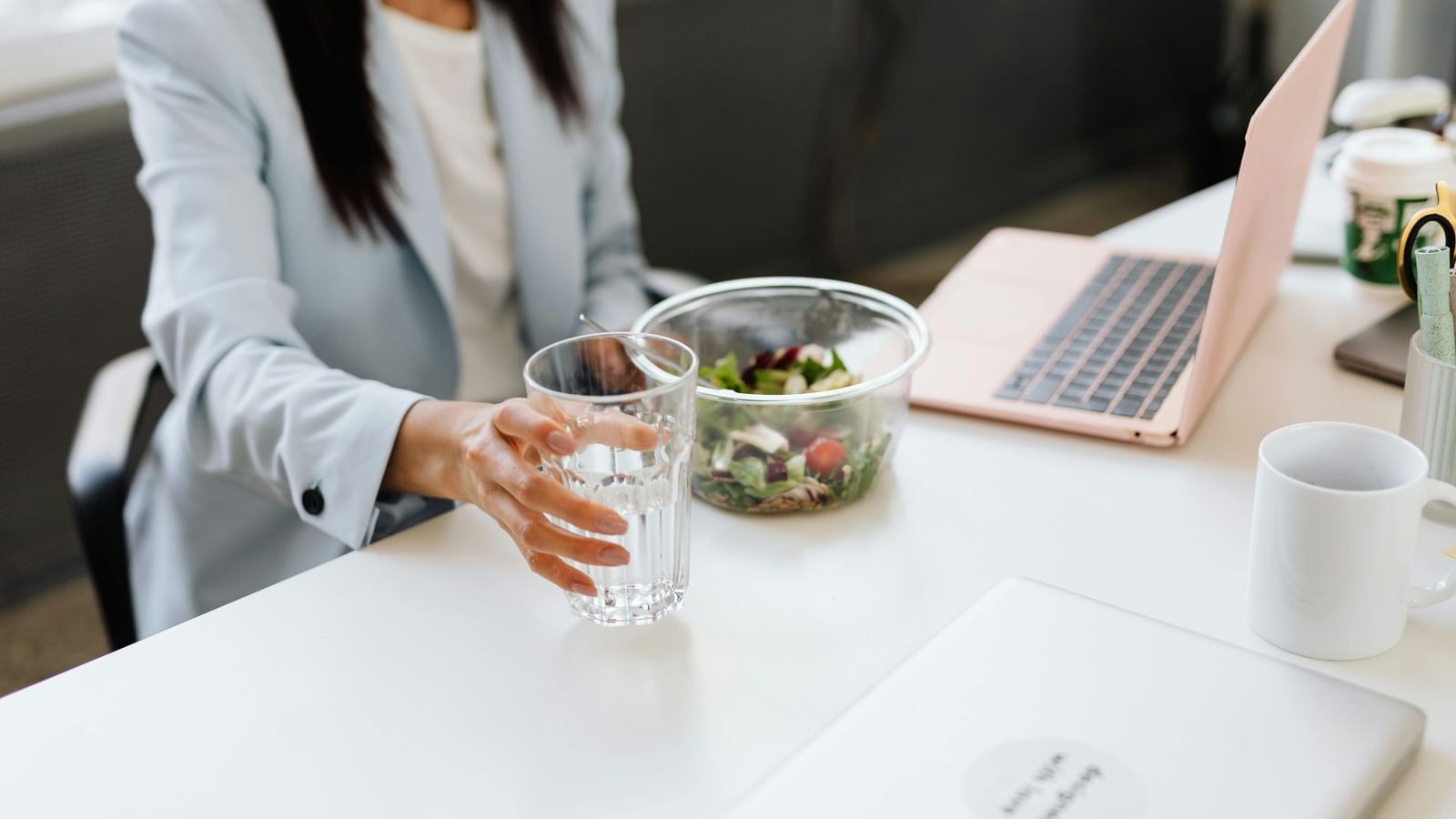 A female employee holding a glass of water, bowl of salad, a laptop and coffee mug on a table