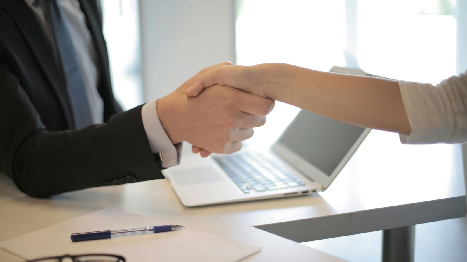 Two people shaking hands, a table with laptop, pen and a pair of glasses