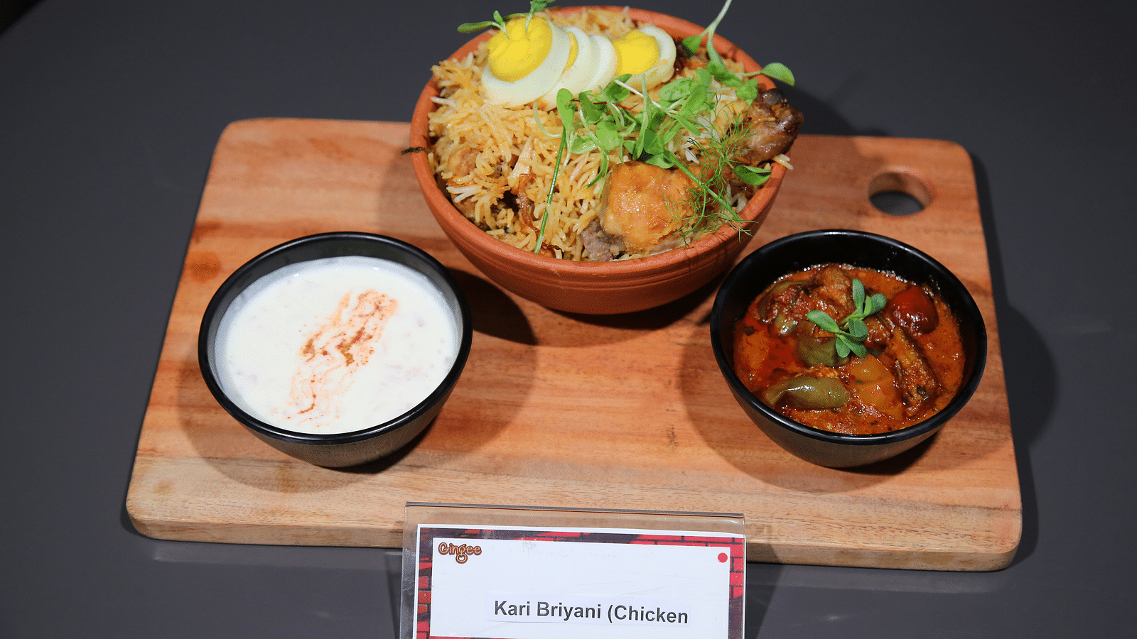 A wooden platter with three bowls. The center bowl contains noodles, half a boiled egg, and garnishes. The left bowl holds a white creamy liquid, and the right bowl contains a reddish curry.