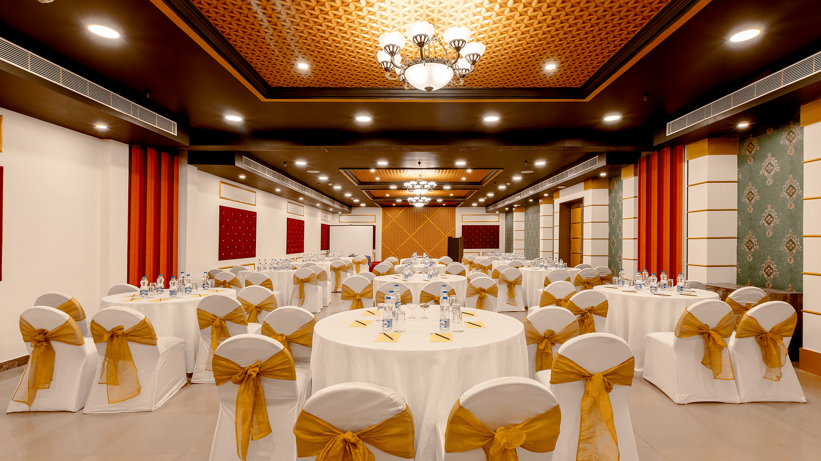 A spacious banquet hall with round tables covered in white cloths and gold chair covers. The hall has a modern design with dark wood accents and a crystal chandelier.