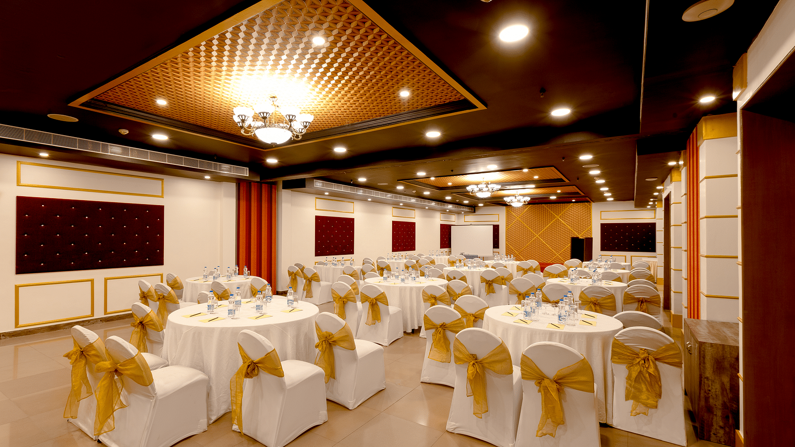 A spacious banquet hall with round tables covered in white cloths and gold chair covers. The hall has a modern design with dark wood accents and a crystal chandelier.