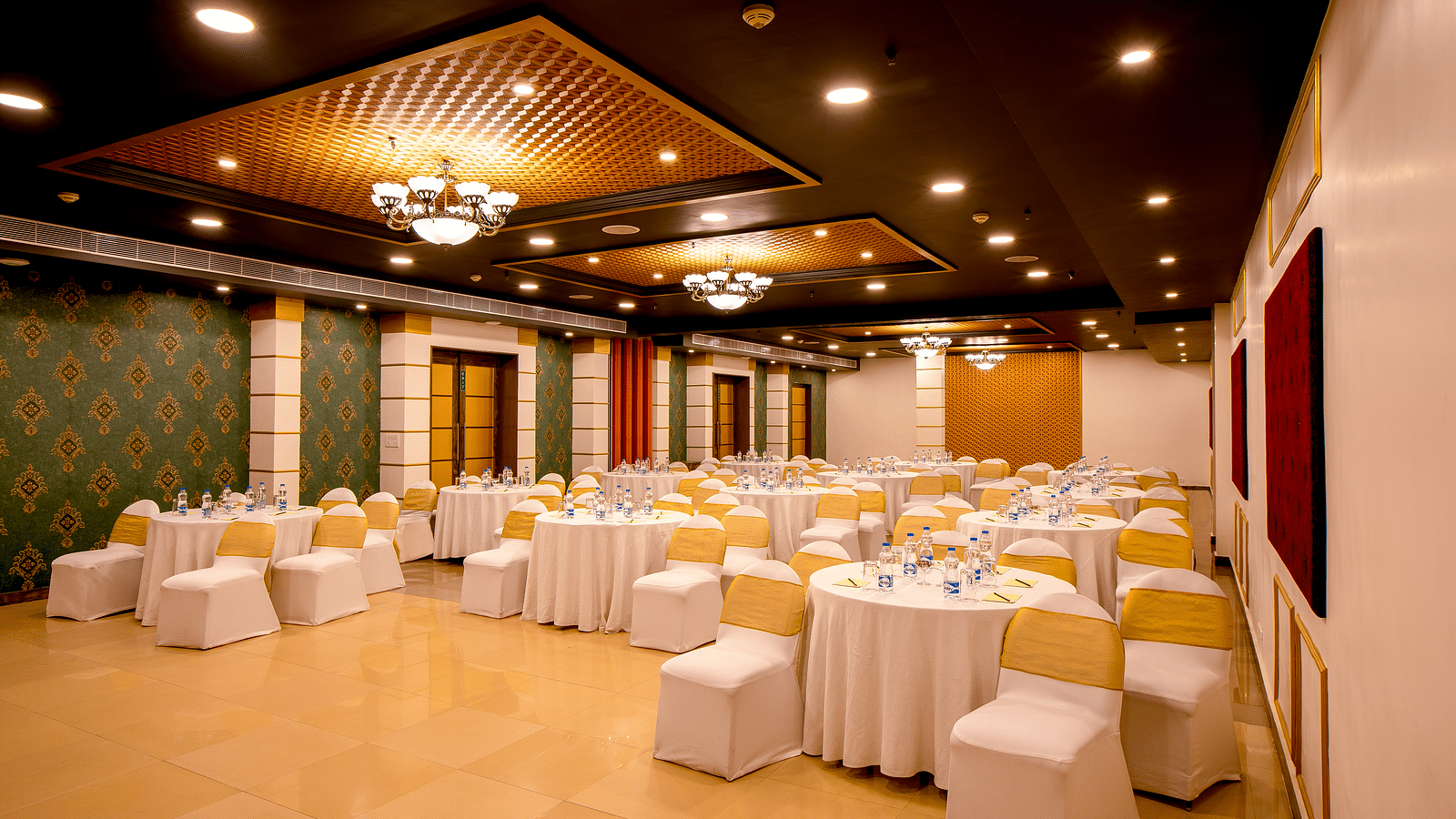 A spacious banquet hall with round tables covered in white cloths and gold chair covers. The hall has a modern design with dark wood accents and a crystal chandelier.