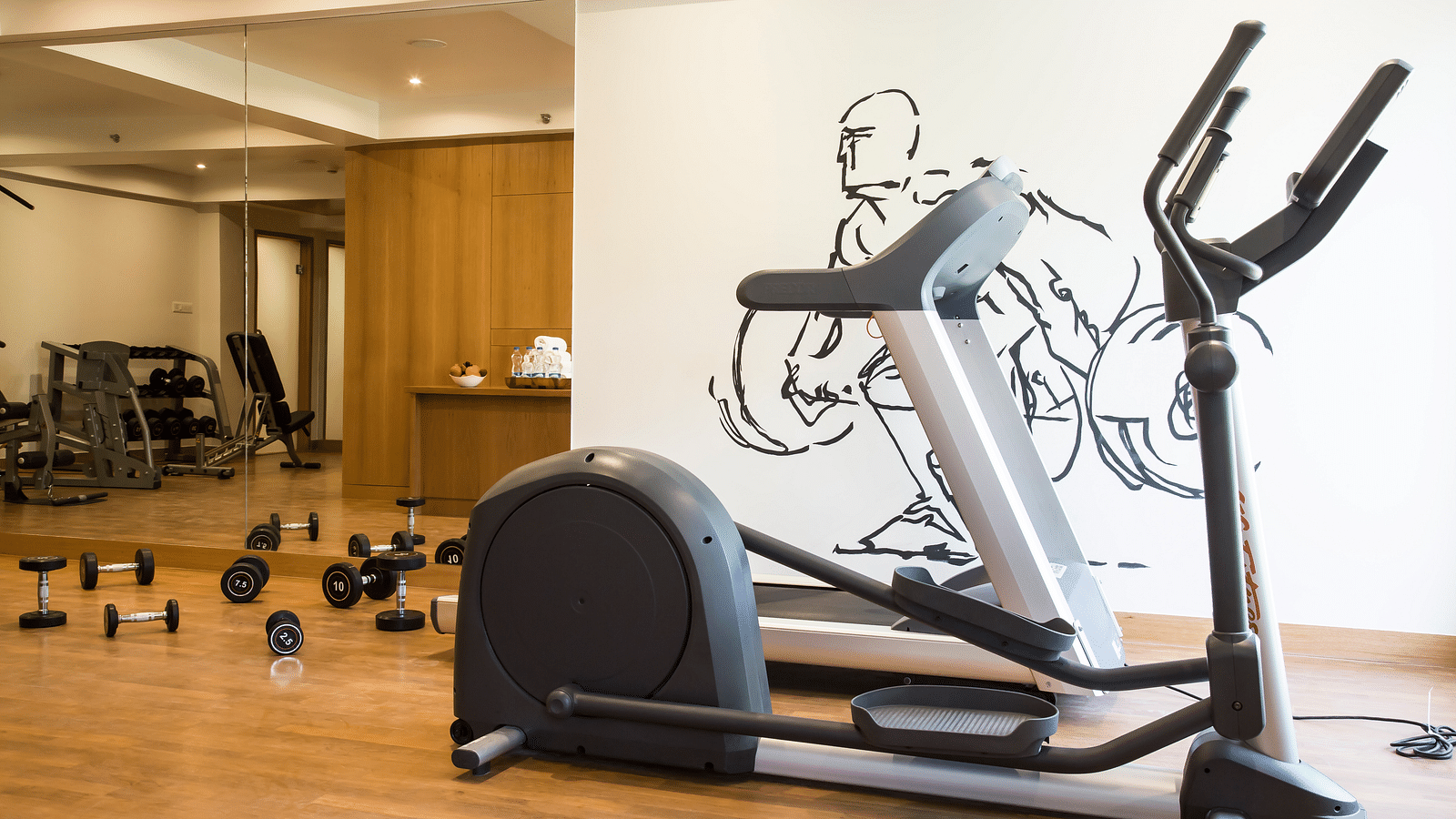 A gym interior with a treadmill, elliptical machine, dumbbells, and a wall mural of a cyclist | Regency Tirunelveli By GRT Hotels.