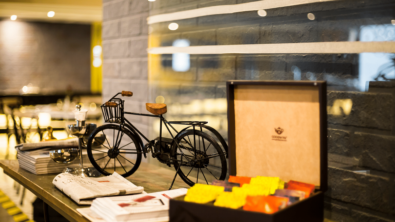 A miniature black bicycle with a basket, next to a wooden box filled with colorful tea packets. The scene is set on a wooden counter in a modern cafe  | Regency Tirunelveli By GRT Hotels