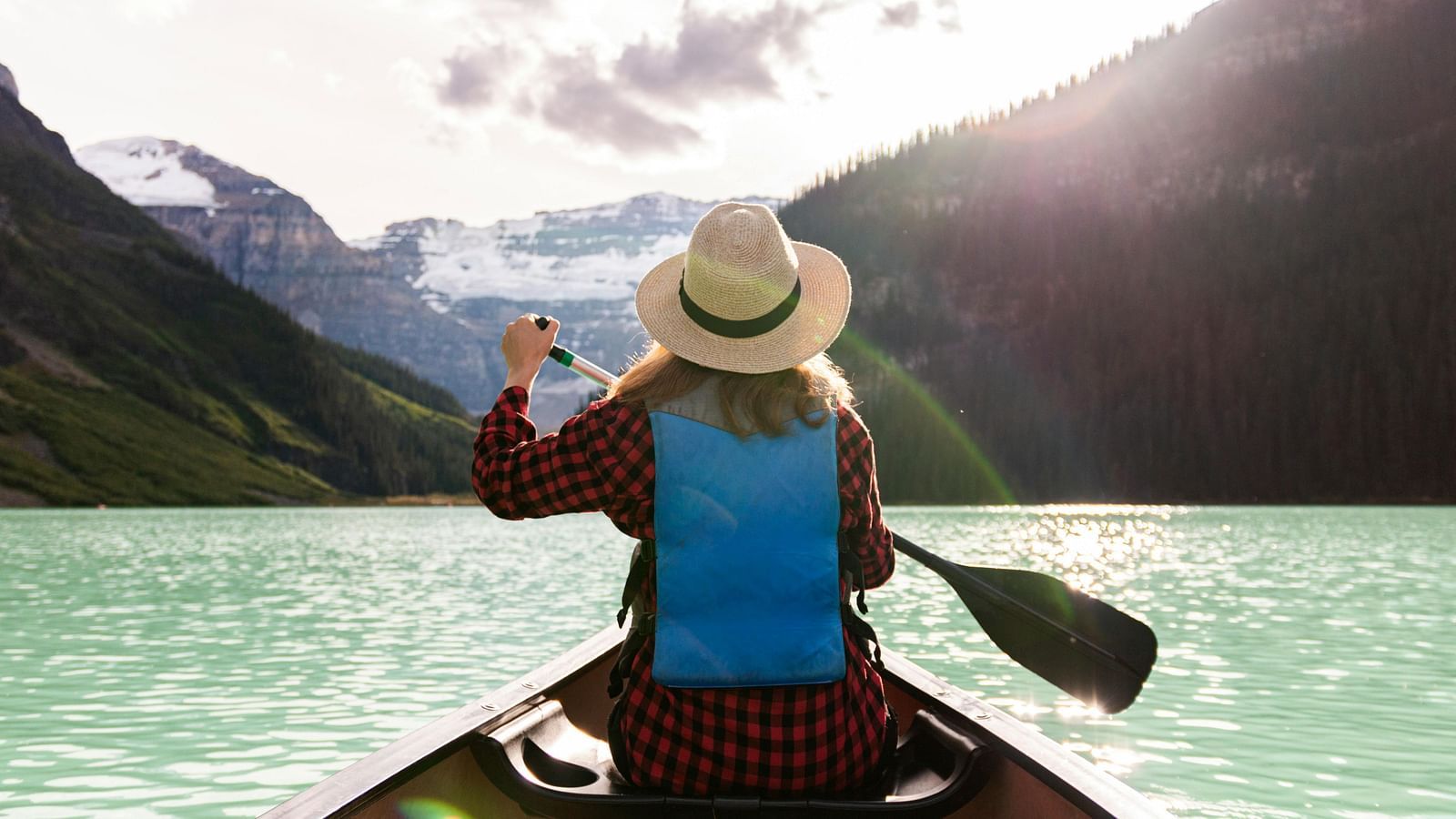 An image of a person in a hat and life jacket is canoeing on a glacial lake, with majestic snow-capped mountains in the background - Kumarakom 