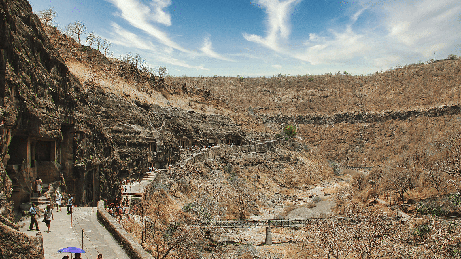 A panoramic view of ancient rock-cut caves against a barren hillside, with visitors walking along a path