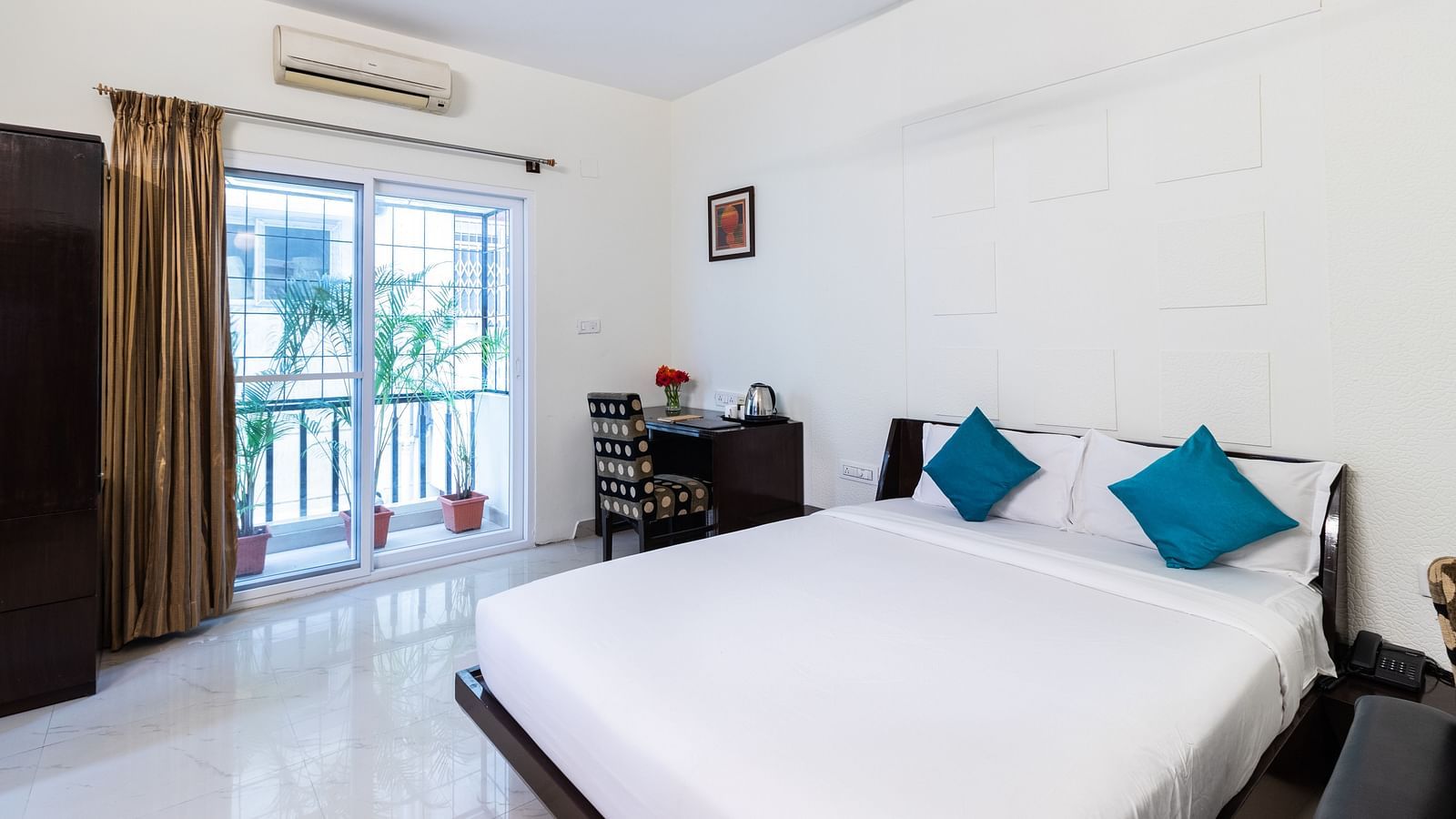 A room with a double bed with blue pillows, a work desk and chair, and glass door leading to the balcony at Sanctum Suites Domlur, Bangalore.
