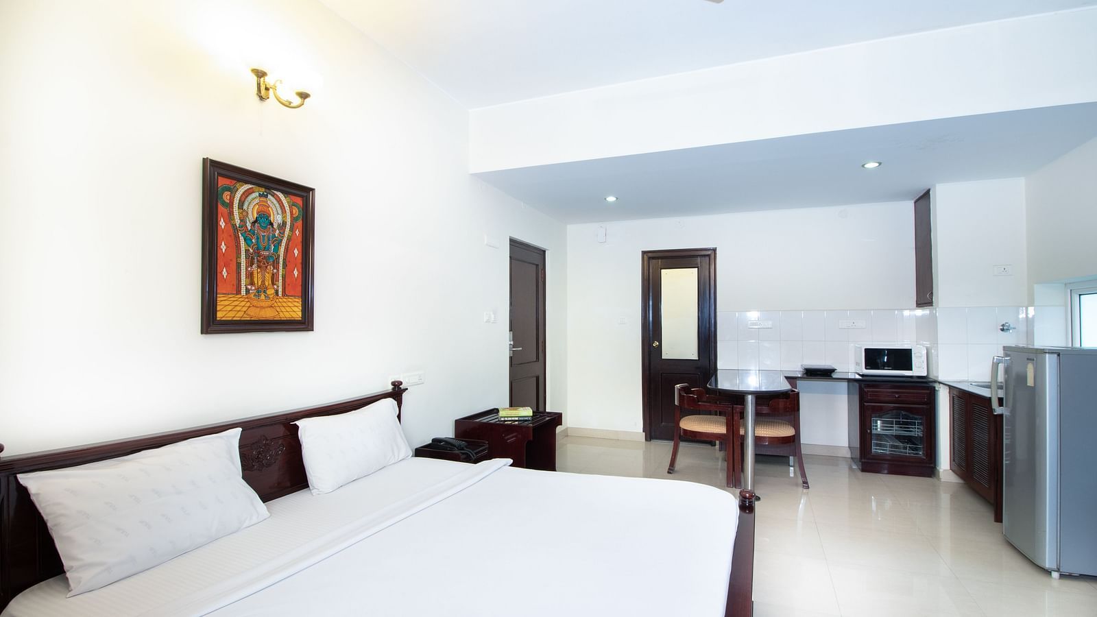 A room with a double bed featuring white linens, a ceiling fan, a framed picture on the wall, a dining counter, a refrigerator, and the kitchenette area at Sanctum Suites Indiranagar, Bangalore.