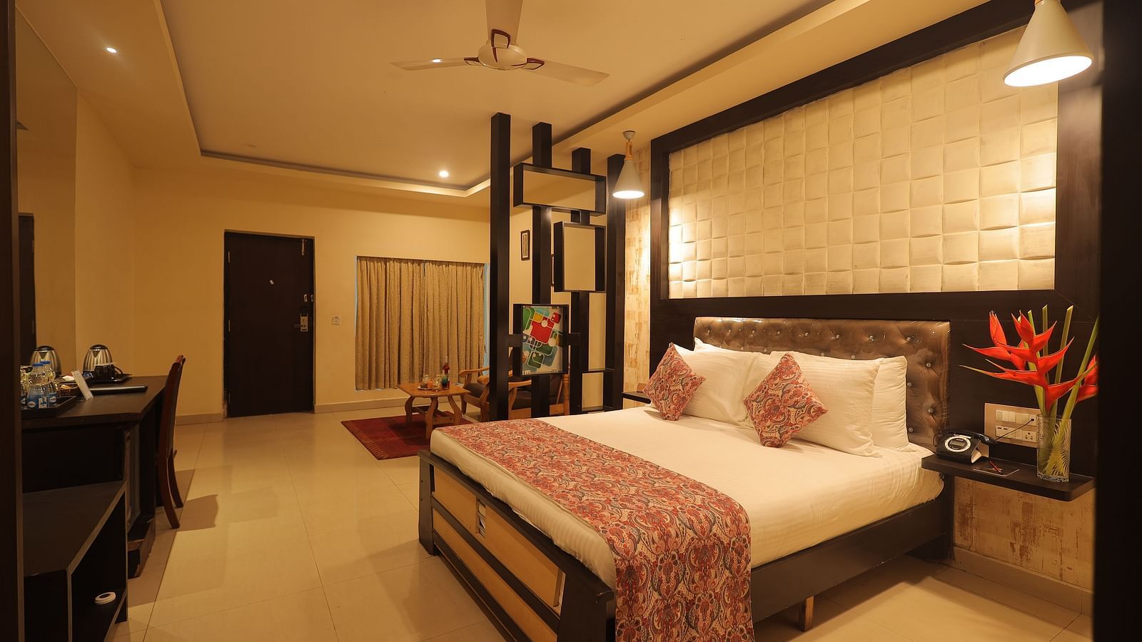 Spacious, warmly lit room at Sathyam Grand Resort with a large double bed, elegant furnishings, decorative cushions, fresh flowers, and a comfy seating area.