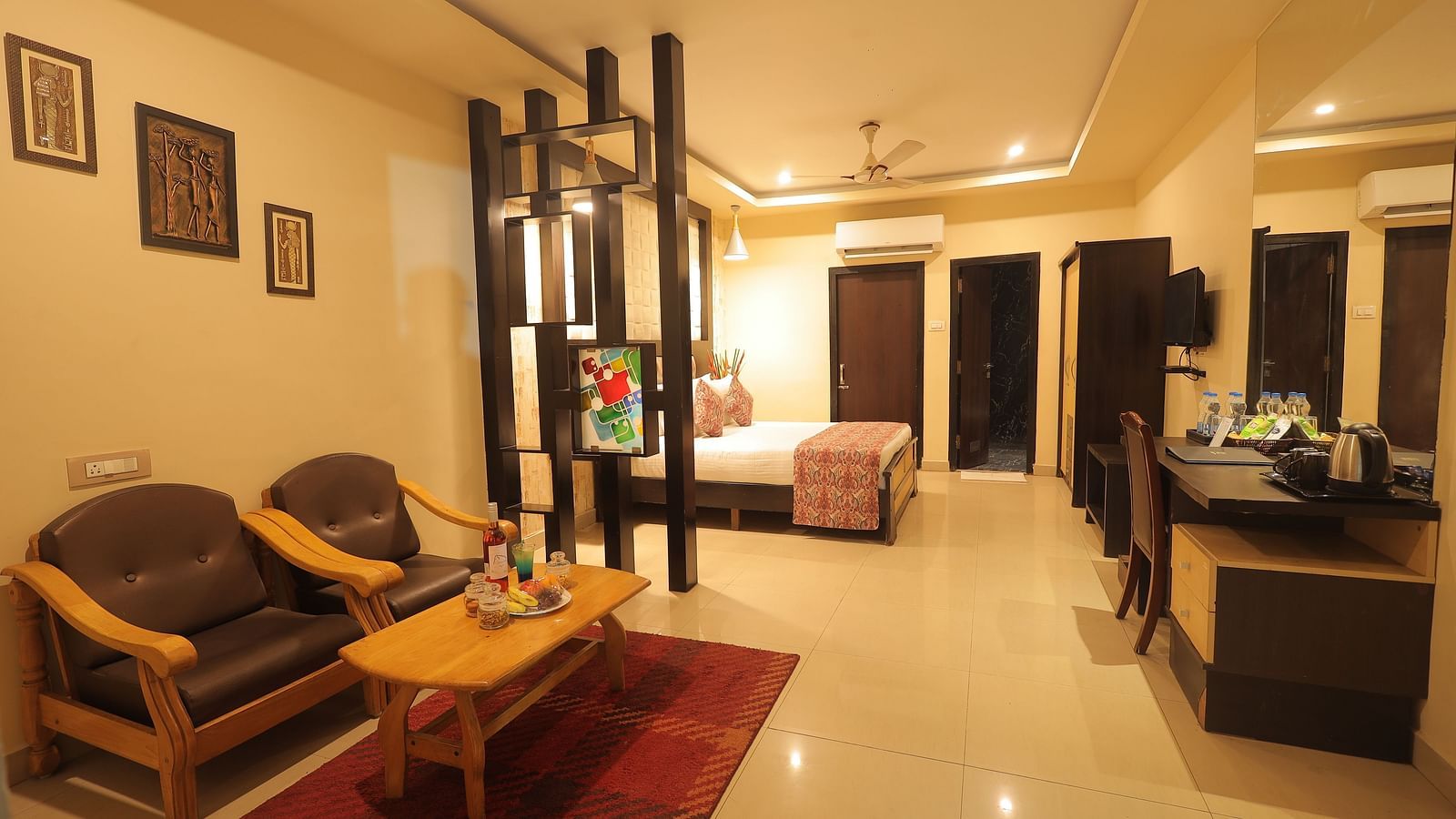 A living space at Sathyam Grand Resort with bedside table, a set of table and chairs, two sofa seats, flat TV screen, and AC
