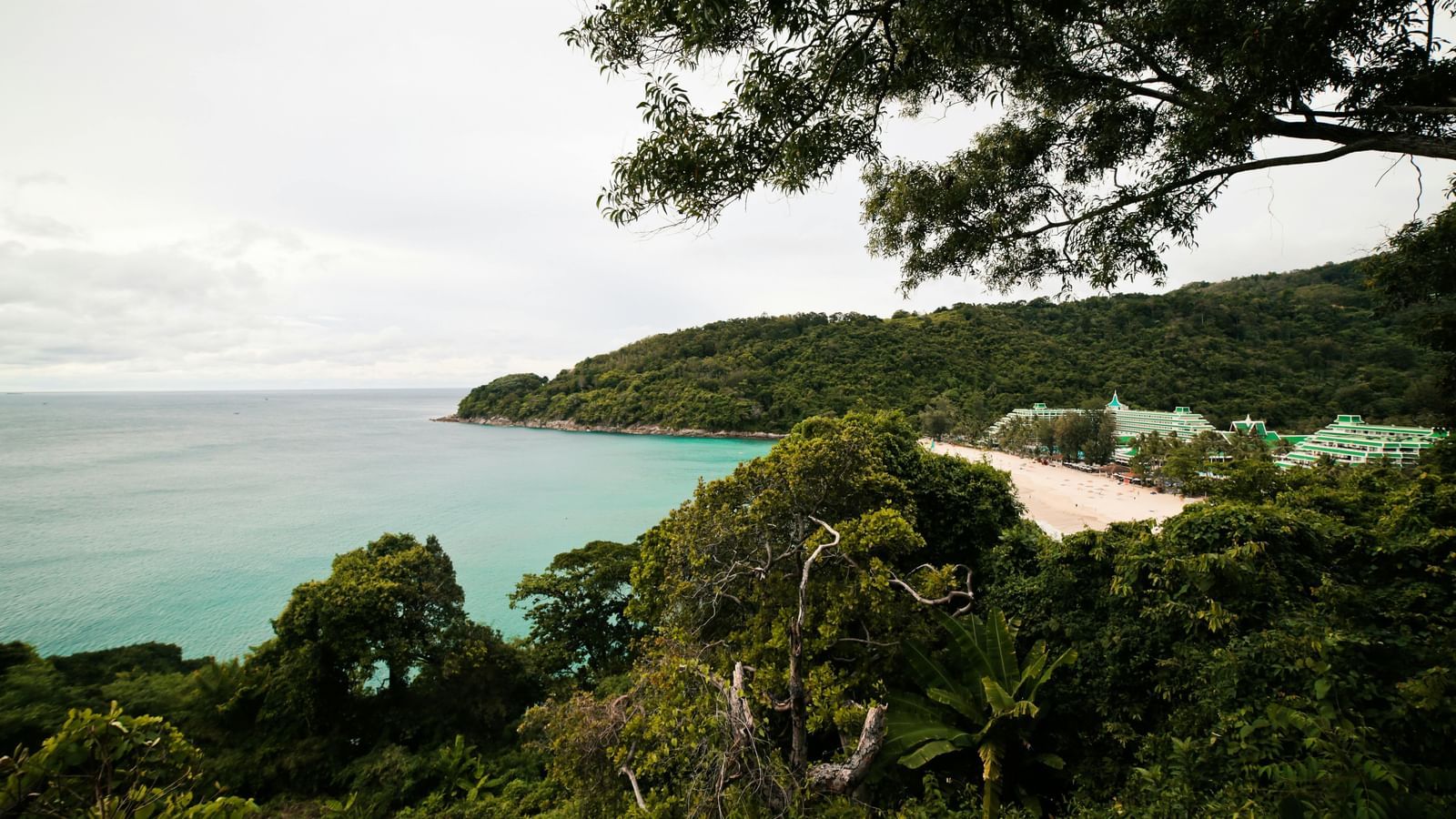 A view of a lush green headland jutting into the clear blue sea, with a cloudy sky in the background.