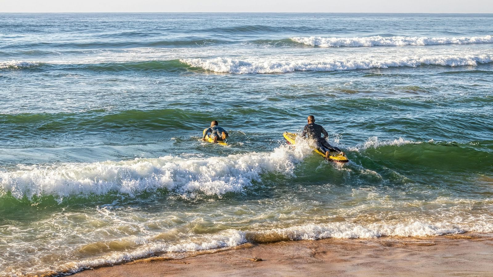 Two people surfing on breaking waves near a sandy beach.