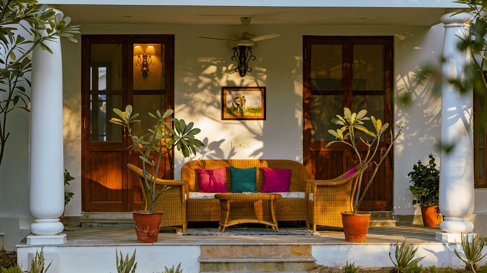 A porch with a wooden sofa and colorful cushions is flanked by two dark wooden doors, with potted plants and white columns on either side- Shahpura Bagh.