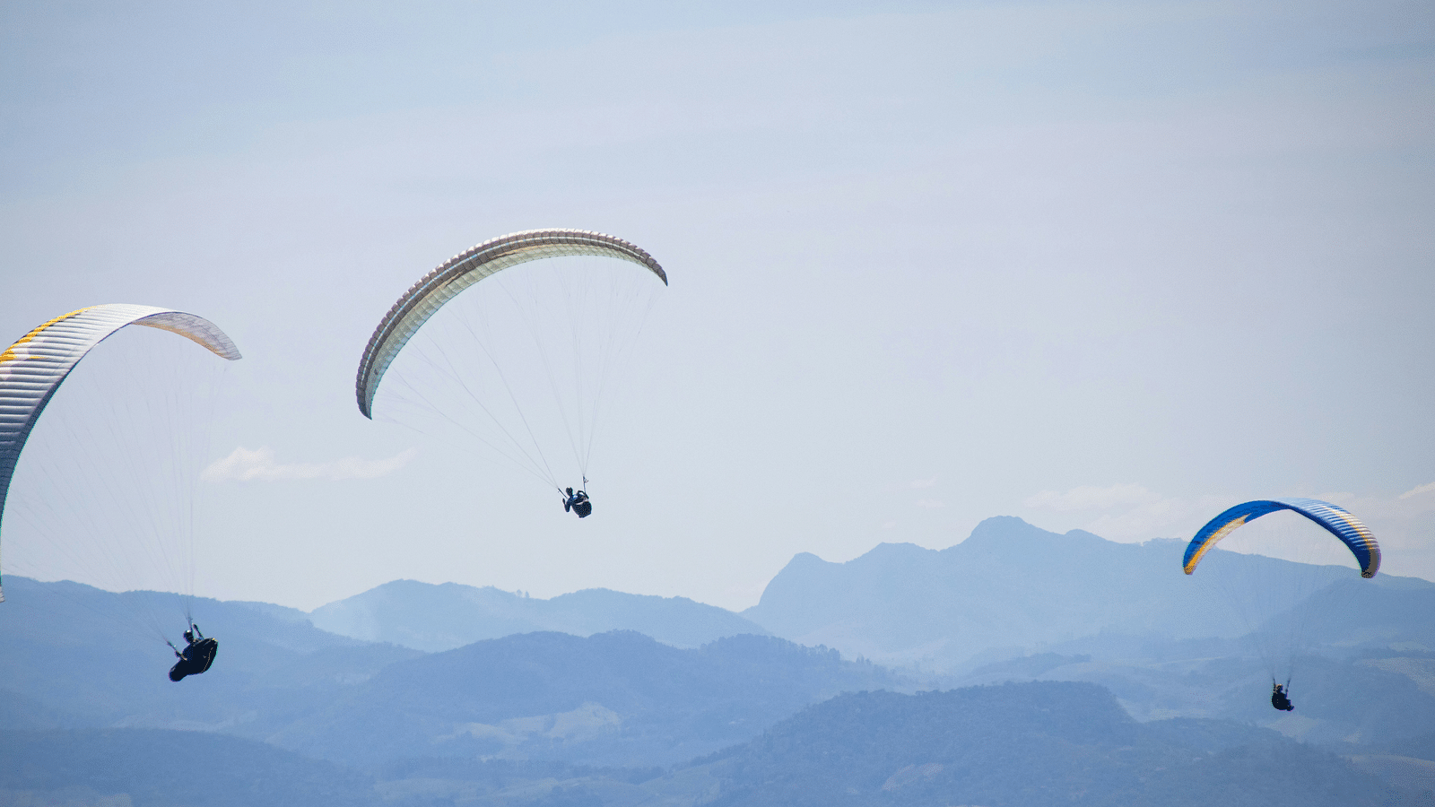 Three people paragliding with different parachutes with many mountains in view.