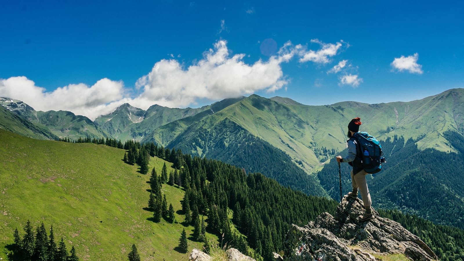 A man standing on the edge of a cliff looking at the mountains on a sunny day.