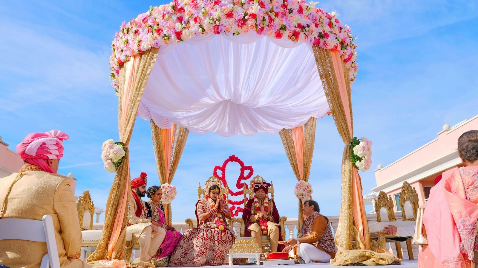 Beautiful outdoor wedding mandap decorated with colourful flowers and drapes, set under a clear blue sky with the couple seated inside.