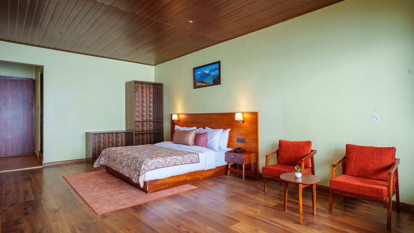 A well-appointed hotel room at Summit Enigma Resort & Spa, Lamahatta, featuring a large bed, two red armchairs, and a warm, inviting ambience.