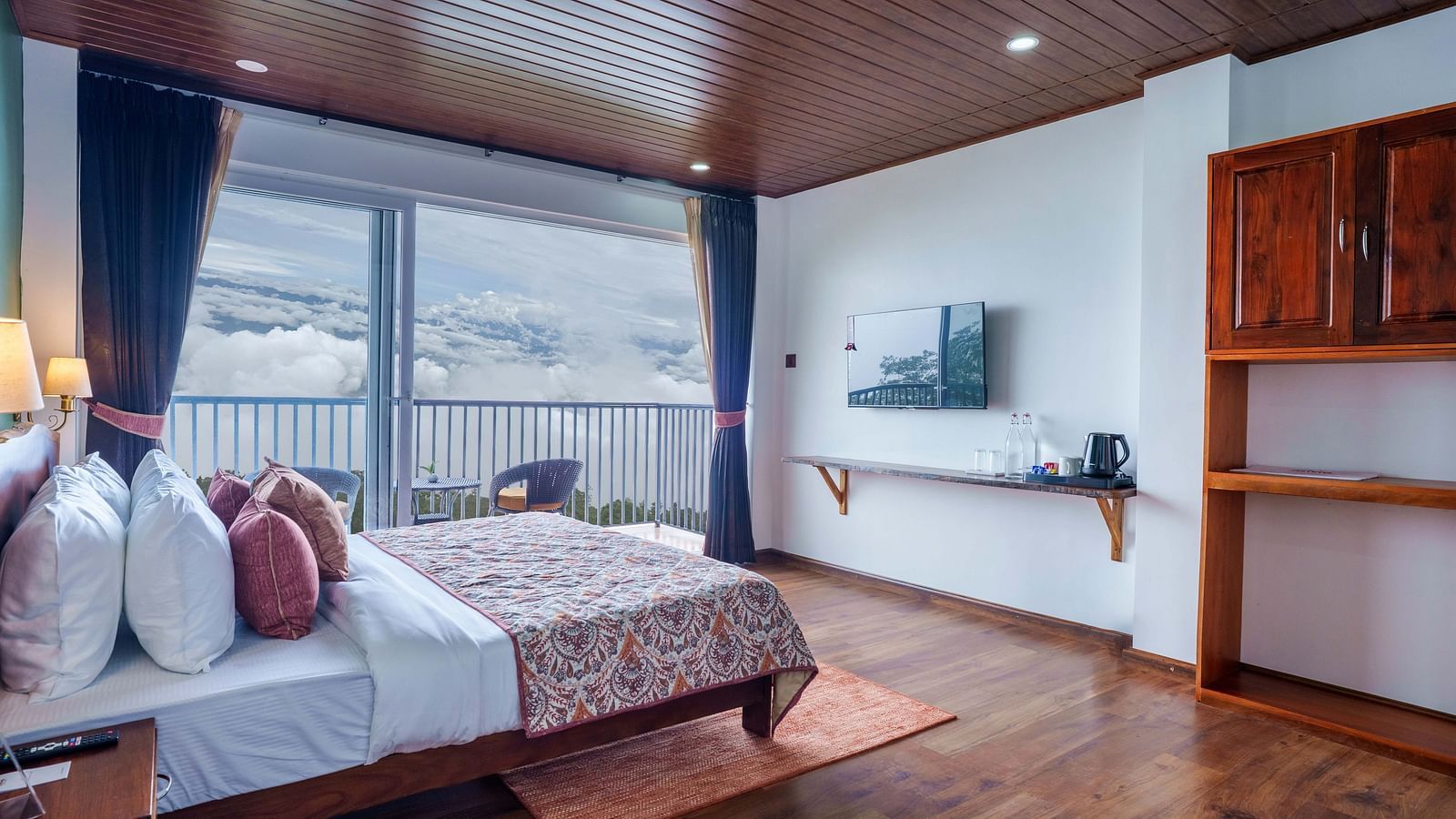 An interior shot of a room at Summit Enigma Resort & Spa, Lamahatta, showcasing a comfortable bed, large windows offering panoramic views, and wooden flooring.